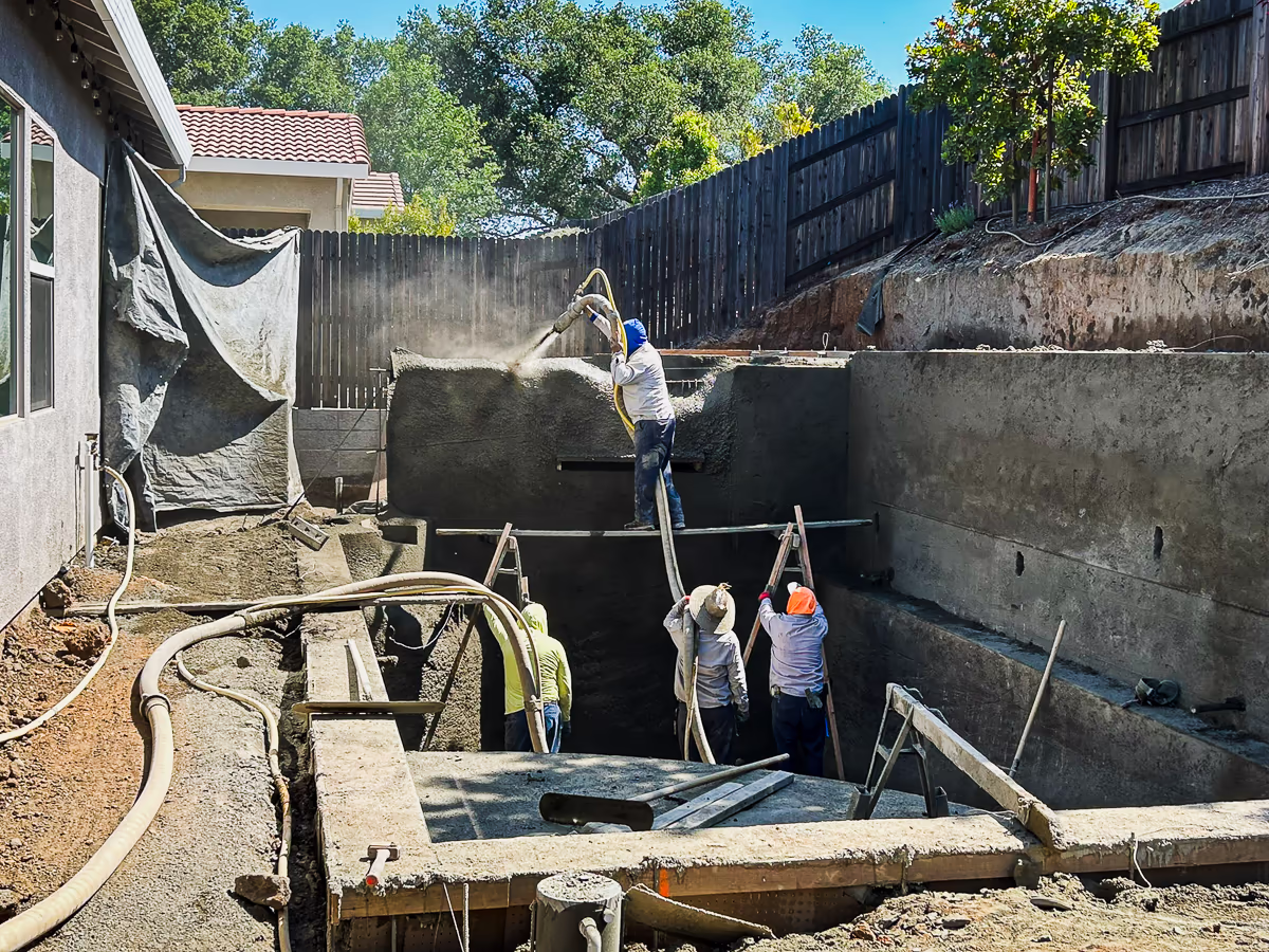 Gunite crew shooting pool shell and structural retaining wall with house and hillside visible during small yard pool construction in Rocklin CA