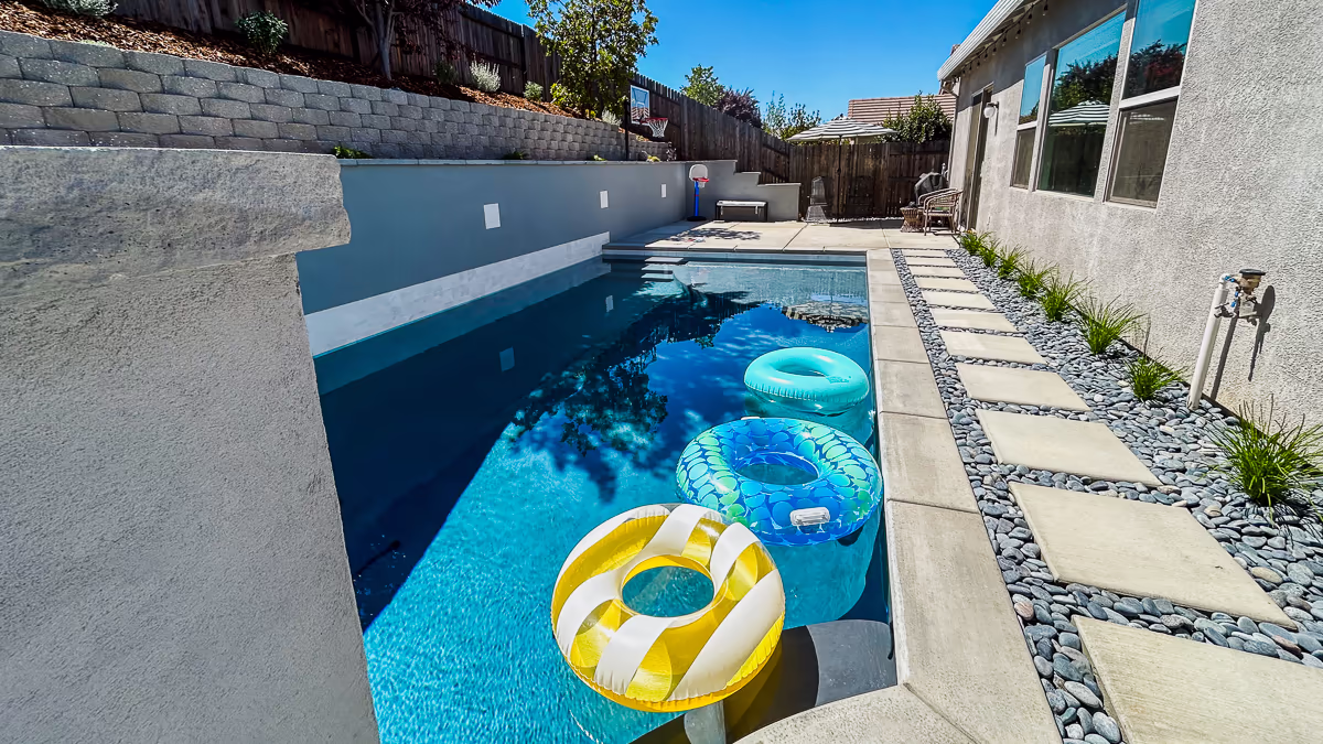 Finished small yard hillside pool in Rocklin California with Tahoe Blue plaster, stepping stone side yard walkway, stacked block upper retaining wall, and pool floats showing family use