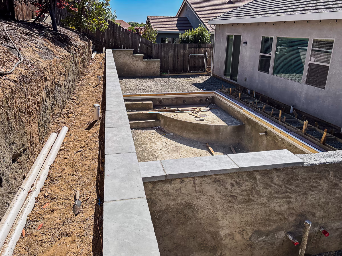 Elevated overview of small yard hillside pool construction in Rocklin California showing completed Eldorado Stone wall cap on gunite retaining wall, pool shell with tanning shelf, deck rebar forms, and hillside drainage pipe