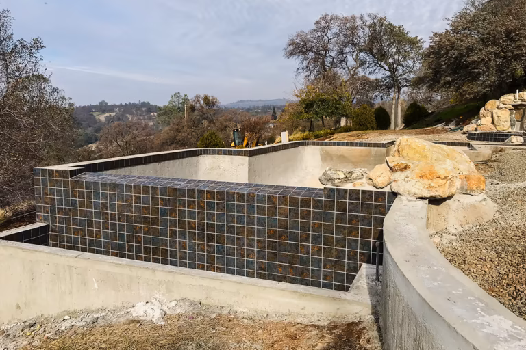 Completed Auburn vanishing edge pool tile weir wall — 36 inches of dark slate tile on face, tiled weir shelf sloping toward catch pool, wading pool and waterfall upper right, Auburn foothill views beyond
